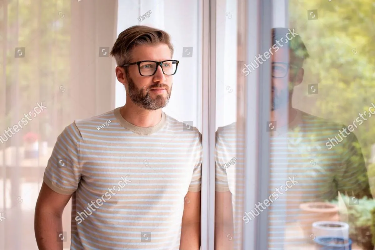 Bearded man in glasses leaning by a window, gazing outside thoughtfully