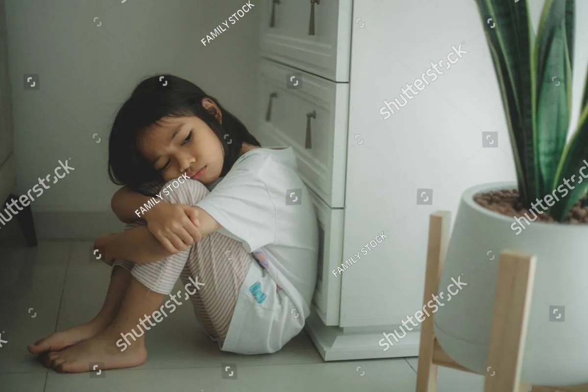 A young girl sits on the floor hugging her knees with her eyes closed