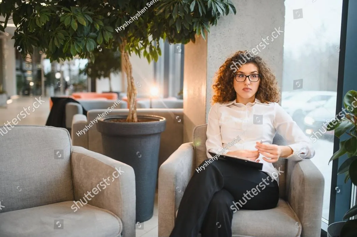 A woman in glasses sits in an armchair with a notebook, listening attentively