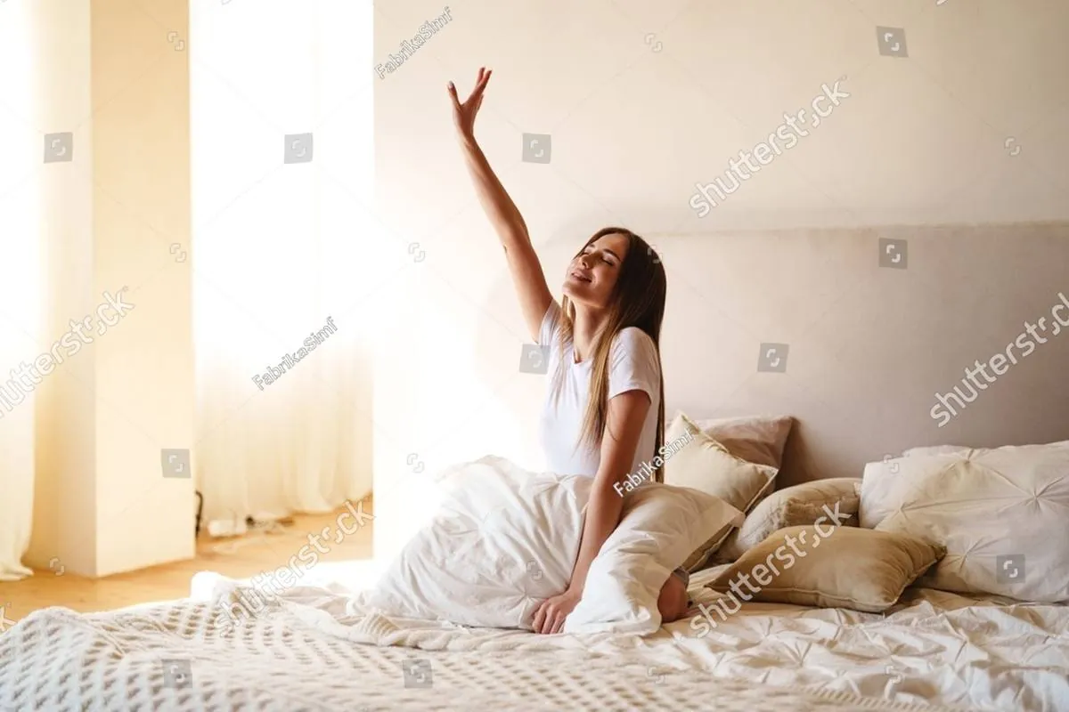 Smiling woman stretching one arm up while sitting in a sunlit bed