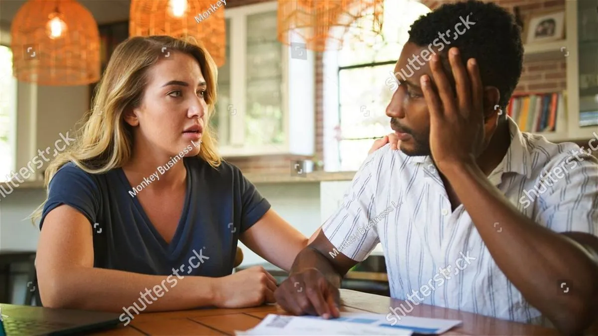 A frustrated couple sits at a table with paperwork, the man holding his head