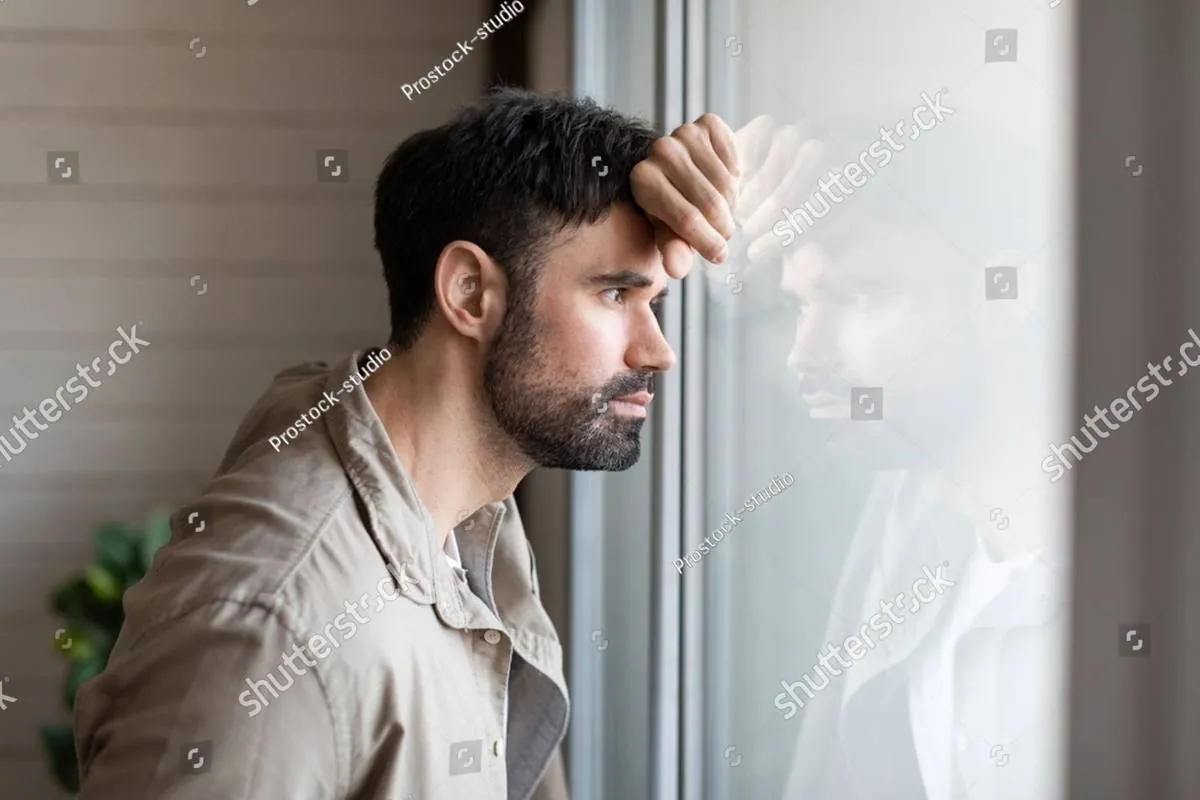 A man leans his forehead on his hand against a window, gazing outside thoughtfully