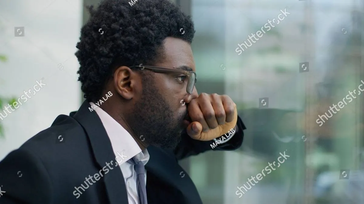 Man in a suit and glasses gazing out an office window, fist to mouth