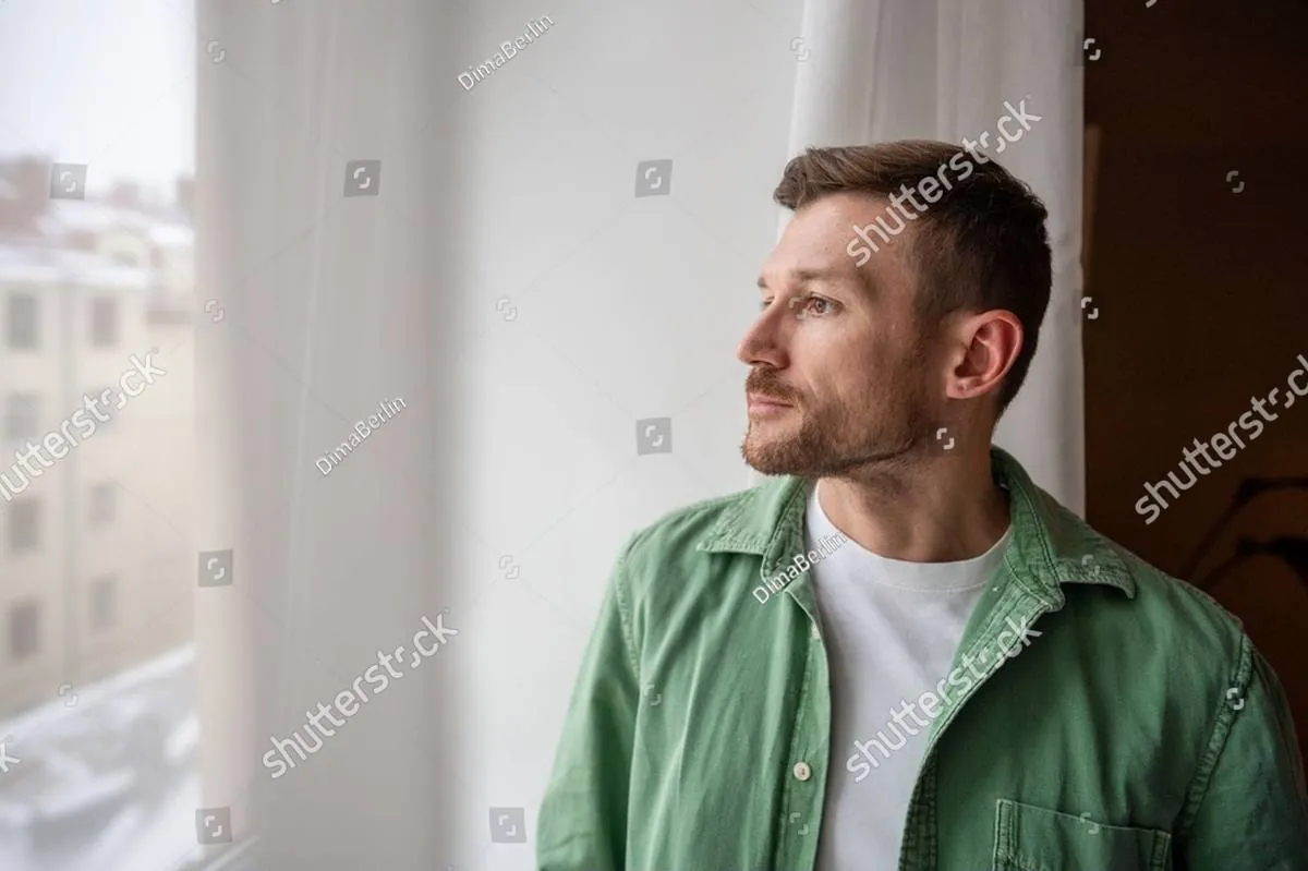 A man in a green shirt looks thoughtfully out a bright window