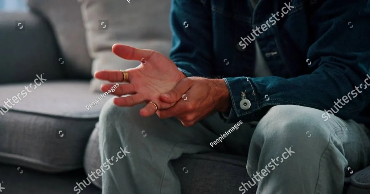 Close-up of a seated man’s hands gesturing as he talks on a gray sofa