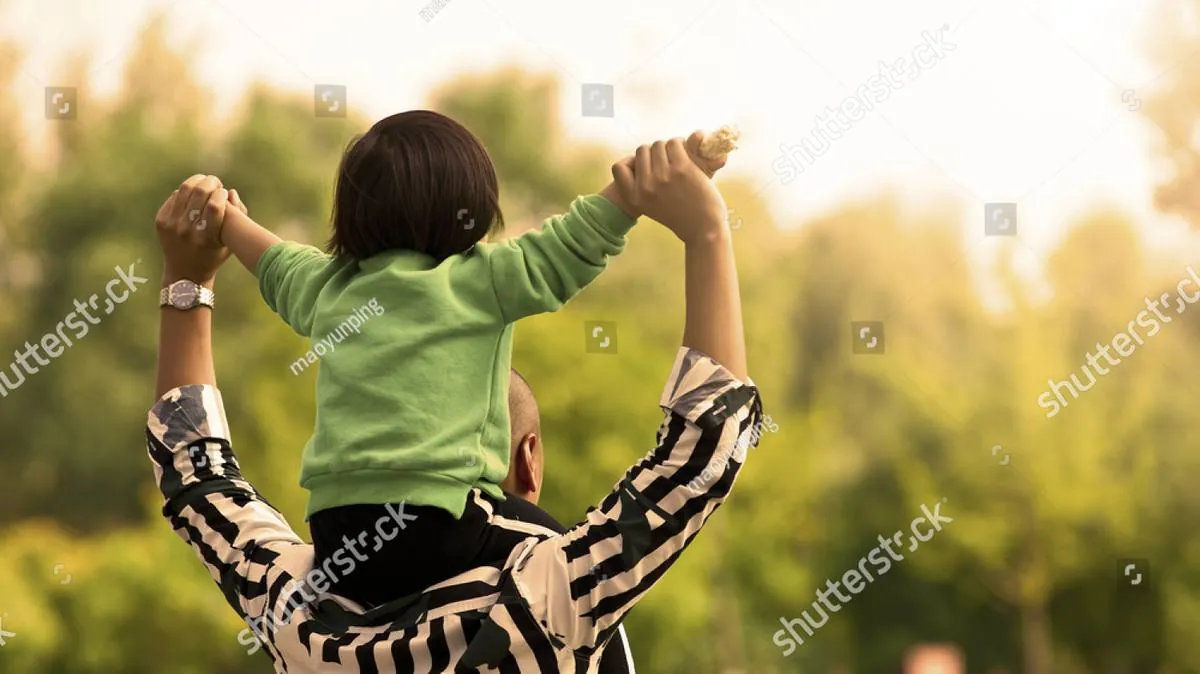 A father carries his child on his shoulders, holding hands raised among green trees