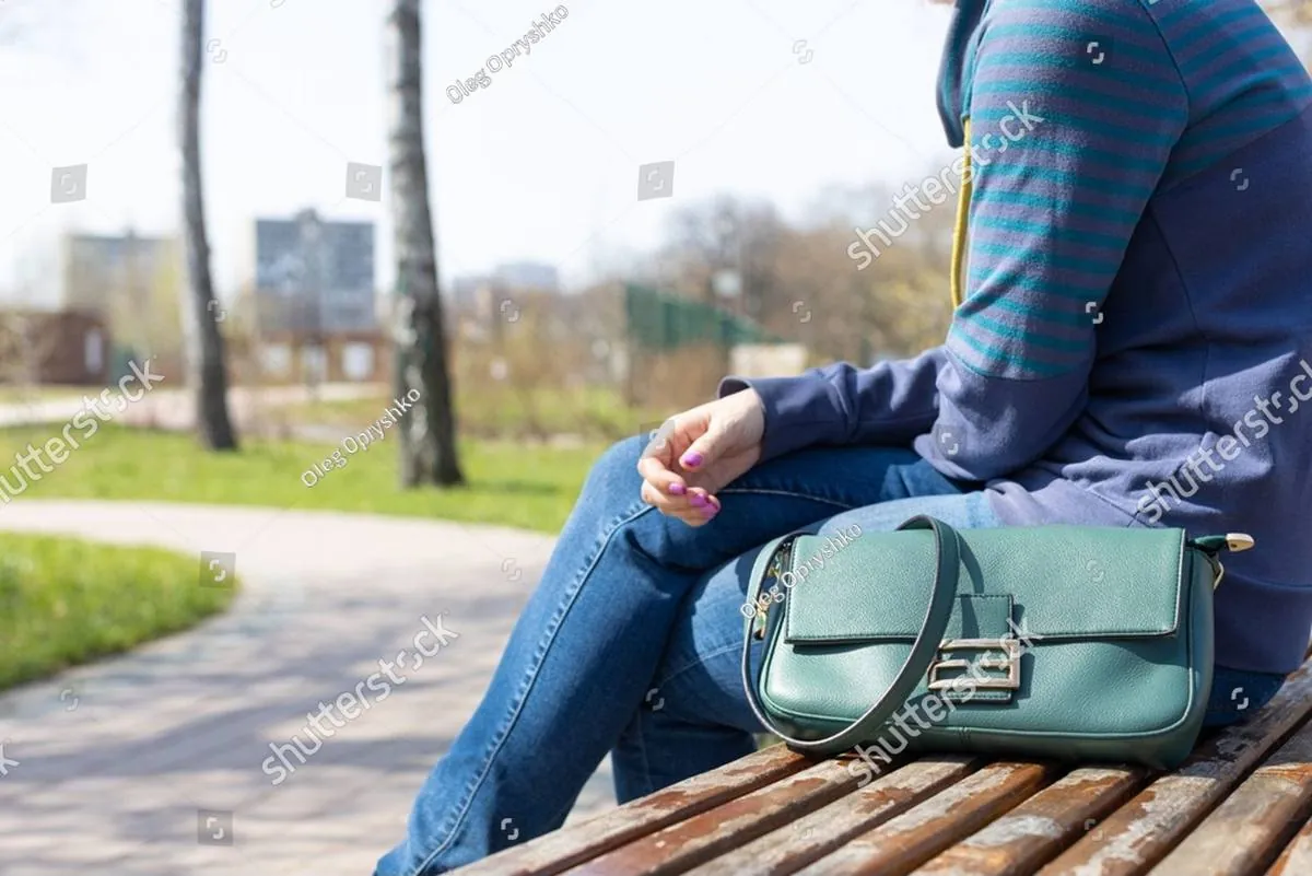 Woman in jeans and a striped sweatshirt sitting on a park bench with a green handbag