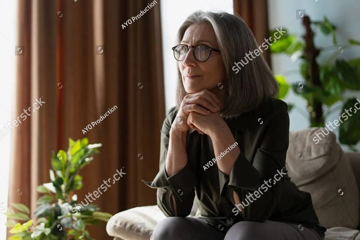 Gray-haired woman in glasses sitting on a sofa, chin resting on her clasped hands