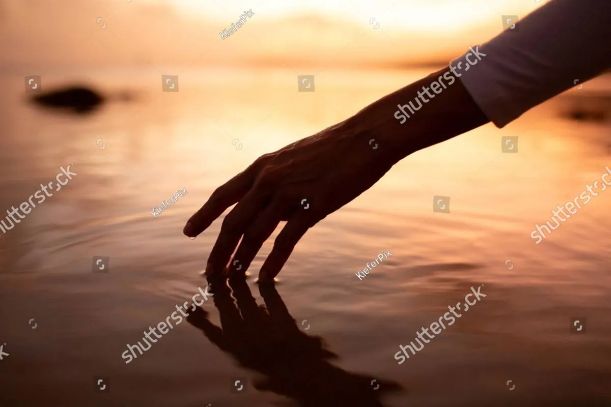 Silhouetted hand touching calm water at sunset, reflected on the surface