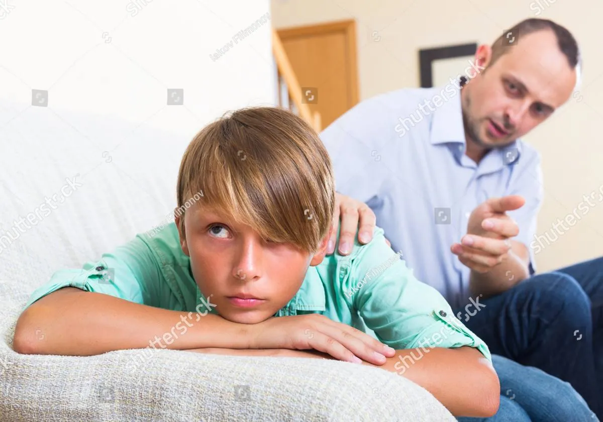 A sullen teenage boy rolls his eyes on the sofa while his father talks to him