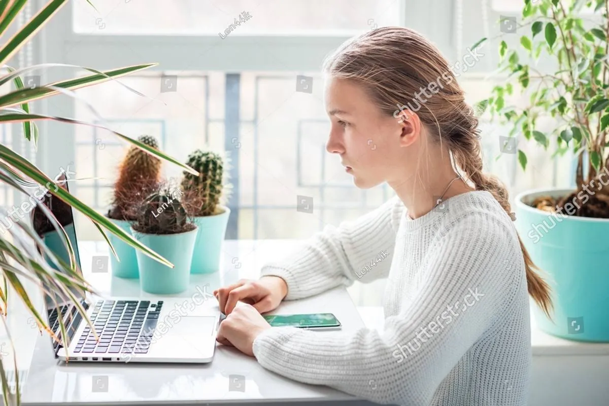 A teenage girl sits at a white desk looking at an open laptop beside potted cacti
