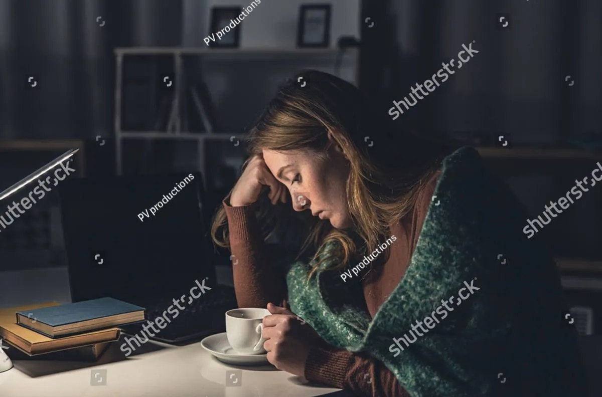 Tired woman wrapped in a green blanket, head in hand at a desk with tea